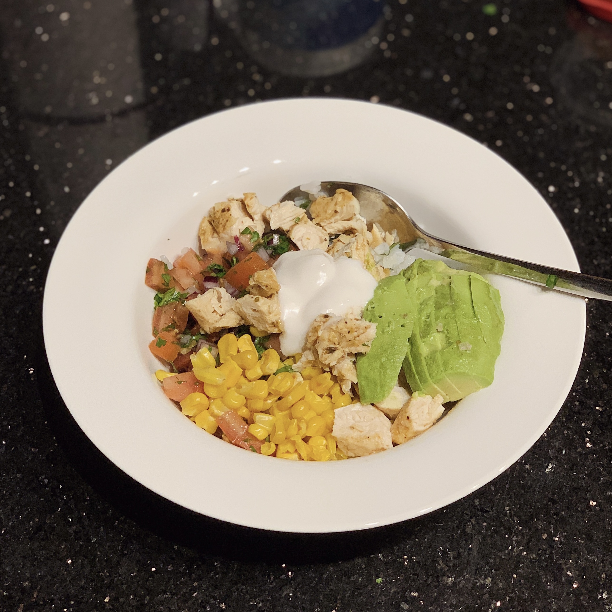 a homemade Chipotle bowl with a spoon, chicken, pico de gallo, corn, avocado, vegan sour cream, and brown rice