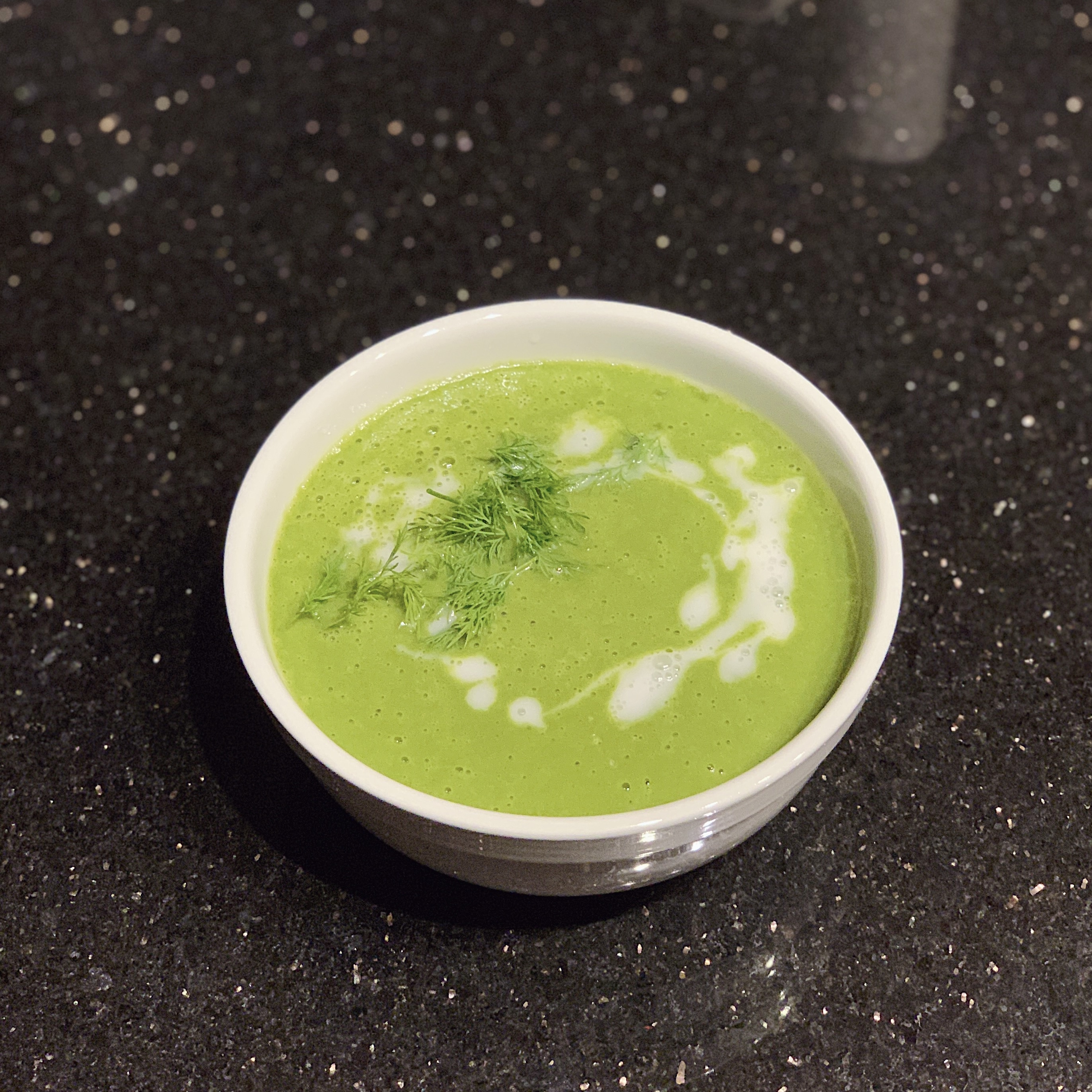 A bowl of Green Goddess Soup garnished with drizzled coconut milk and fresh dill on top.
