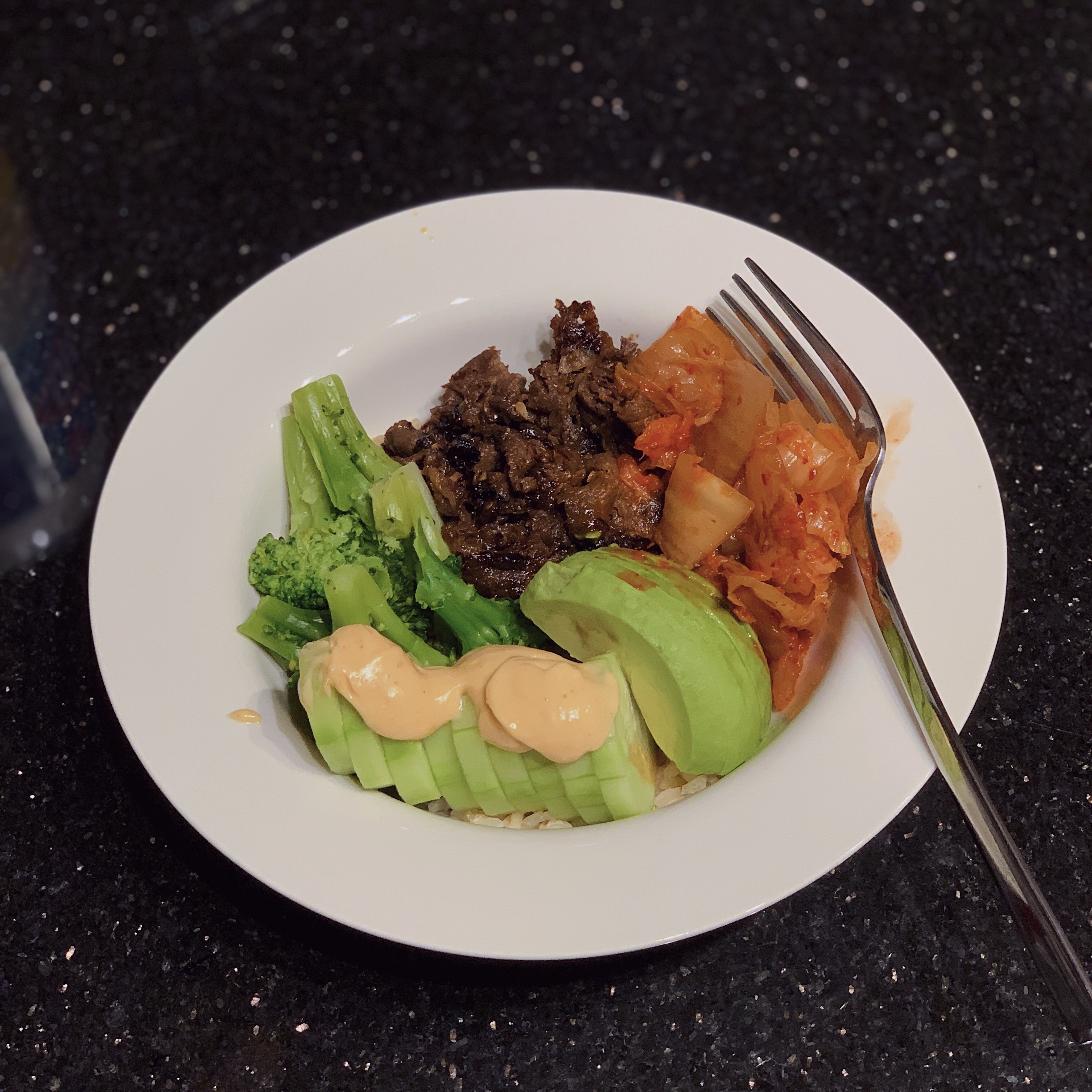 A bowl with kimchi, beef brisket, broccoli, avocado, cucumber and brown rice.